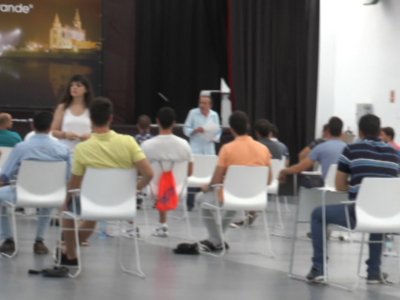 Una sala de conferencias con una pantalla grande en el fondo que muestra un edificio histórico. La audiencia está sentada en sillas blancas, mirando hacia adelante. Algunos asistentes están escribiendo en sus portátiles o tomando notas. La sala tiene una iluminación blanca y la pared del fondo es de color blanco.