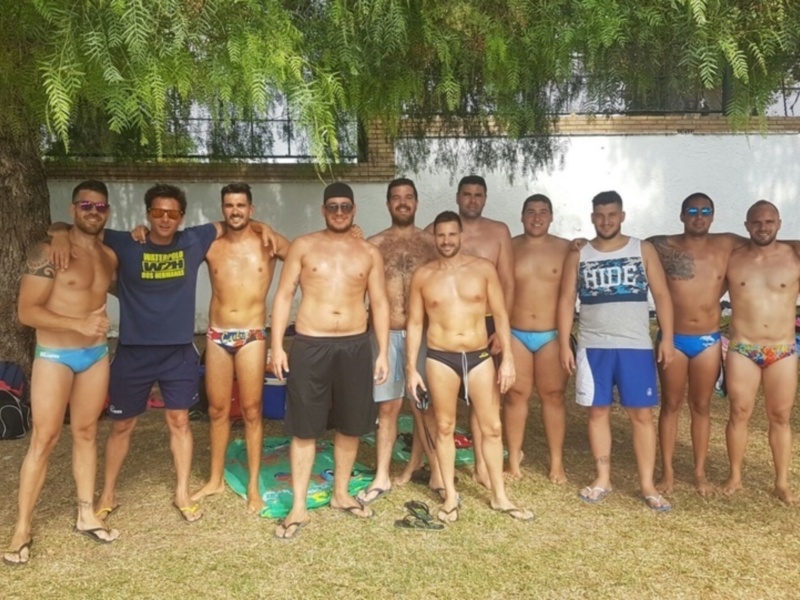 Un grupo de hombres posando en un área al aire libre, vestidos con ropa de playa. Algunos llevan pantalones cortos y otros camisetas, con un fondo de vegetación y una estructura blanca.