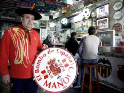 En un ambiente festivo, un hombre con sombrero y camiseta roja sostiene una gran bandera de España. Detrás, se observa un ambiente animado con mesas y decoraciones festivas.