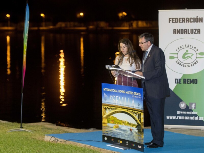 En la imagen, se puede ver a un hombre y una mujer en una presentación frente al agua nocturna. El hombre está sosteniendo un micrófono, mientras la mujer se encuentra detrás de una pantalla que muestra el título "International Synchrony Master Regatta" con la ciudad de Sevilla en el fondo. En el fondo, hay una bandera y un cartel que dice "Federación Andaluza de Remo". La escena se ilumina con luces que reflejan en el agua.