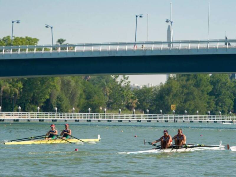 Tres personas remando en una canoa amarilla por el río, bajo un puente. La imagen muestra un día soleado con árboles y una carretera en el fondo.