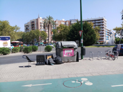 En un entorno urbano, se observa una calle con dos bicicletas aparcadas cerca de un poste. Al fondo, se pueden ver edificios con balcones y árboles. En el lado izquierdo, hay un camión de reparto y varios coches estacionados. En el centro, se encuentra una papelería con un cartel rosa y negro. La imagen muestra una combinación de arquitectura urbana, transporte público y elementos de la vida cotidiana.