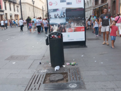 Se observa un callejón con gente caminando y una gran publicidad en el fondo. En la parte inferior de la imagen hay un contenedor de basura lleno de desechos.