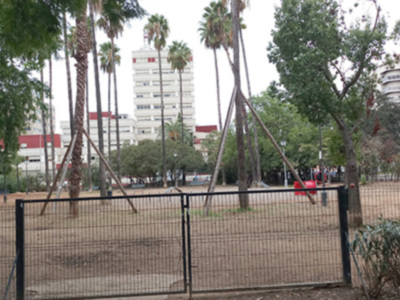 Un parque infantil con columpios y un área de juegos, rodeado por árboles altos y edificios en el fondo.