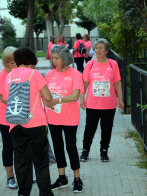 Una imagen de varias personas vestidas con camisetas rosas y pantalones negros, posiblemente participantes de una carrera o actividad física al aire libre. La escena se desarrolla en un entorno urbano con árboles y una valla de metal.