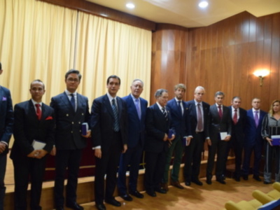 Una fila de personas vestidas con trajes formales posan para una foto en un salón de conferencias con cortinas amarillas y una pared de madera.