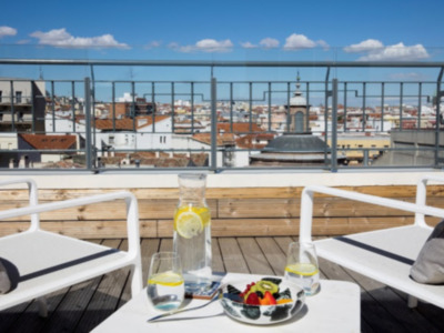 Una terraza con una mesa de madera y sillas blancas, en la que se puede ver un vaso con limón, una jarra de agua y un plato con frutas. En el fondo se puede ver una ciudad con edificios de varios pisos y un rascacielos. El cielo es azul con algunas nubes blancas.