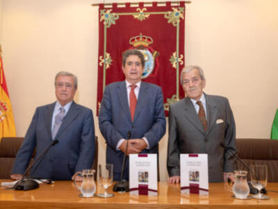 Tres hombres en trajes formales posan frente a una bandera española y otra regional, con un fondo blanco. En el centro, un hombre sostiene una hoja de papel y dos hombres a sus lados tienen portafolios con documentos. La imagen sugiere una reunión oficial o conferencia de prensa.