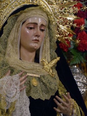 Una figura religiosa vestida en trajes de alta costura, con un atuendo dorado y blanco que incluye una capa con bordados y un tocado elaborado. La figura tiene una expresión serena y está rodeada de flores rojas, lo que sugiere un contexto festivo o ceremonial.