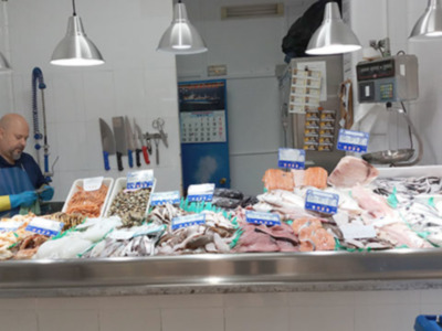 Se ve una escena de un supermercado o tienda de pescados y mariscos, con un mostrador lleno de variedad de productos frescos. En el fondo, se puede ver a un hombre trabajando en la cocina detrás del mostrador. La decoración incluye una pared con un calendario y varios utensilios de cocina colgados. La iluminación es adecuada para una tienda, con lámparas de techo y un reflejante en la parte superior del mostrador.
