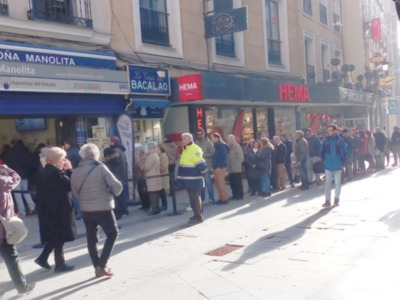 Un grupo de personas esperando en una fila frente a varios negocios en una calle de la ciudad, incluyendo una tienda de loterías y otro establecimiento comercial.