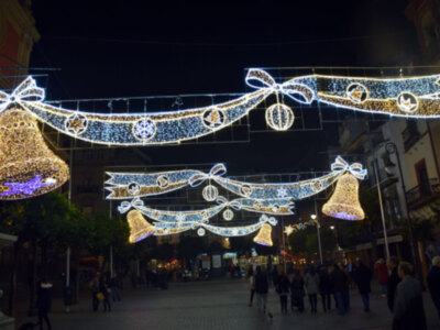 Imagen de una calle iluminada con adornos navideños, incluyendo campanas y cintas de luces doradas y azules. Personas caminando en el fondo, ambiente festivo nocturno.