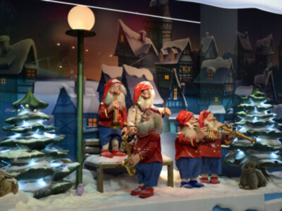 Un grupo de personajes navideños, incluyendo a Papá Noel y otros personajes, se encuentran en un entorno festivo con árboles de navidad iluminados y casas nevadas en el fondo. La escena transmite una atmósfera navideña y festiva.