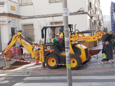 Una escavadora amarilla trabaja en una calle de construcción, rodeada por cinta de seguridad y con casas al fondo.