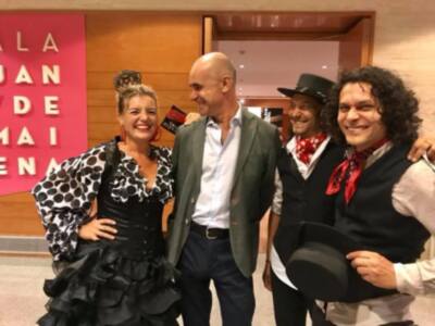 Una mujer viste un vestido negro con lunares blancos, sonriendo junto a tres hombres. Uno lleva un traje verde y sombrero negro, otro tiene una camisa blanca con chaleco negro y corbata roja, y el tercero una camisa blanca con chaleco negro y corbata roja. Al fondo, se ve una pared con letras grandes que forman "MÁLAGA".