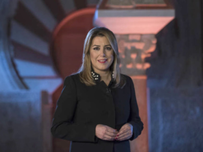 Mujer con chaqueta negra y collar de perlas, sonriendo en un entorno interior con luz suave y detalles arquitectónicos en el fondo.