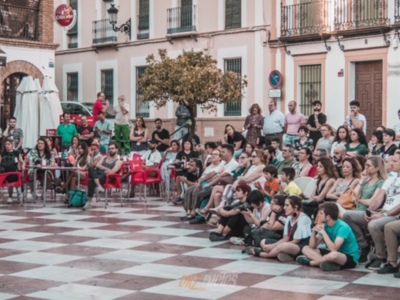 Una multitud de personas se reúne en una plaza, disfrutando de una actuación en el centro. La arquitectura colonial y los locales de comida en el fondo completan la escena.