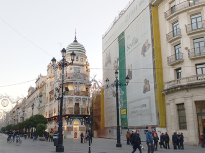 Imagen de una calle con edificios históricos y grandes carteles publicitarios. La arquitectura es clásica con detalles ornamentales y balcones. El cielo está nublado, lo que añade un tono suave a la escena.