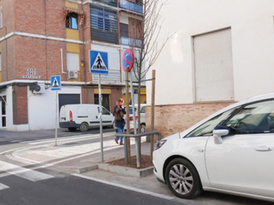 Calle urbana con señal de paso de peatones y zona de estacionamiento regulado, edificios en el fondo.