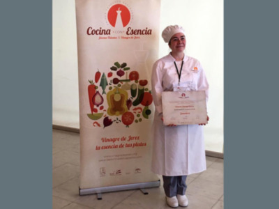A una persona vestida con uniforme de chef, posando orgullosamente frente a un banner que dice "Cocina con Esencia" y presenta un certificado. La escena se centra en la celebración de logros culinarios, enfocándose en el arte y la excelencia en la cocina.
