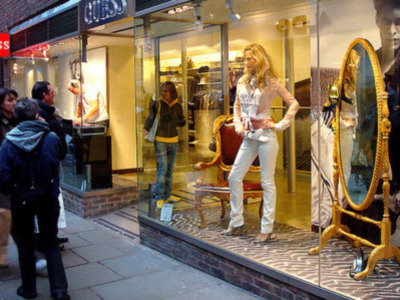 Una mujer con ropa de marca GUESS mira el escaparate de una tienda. Detalles del escaparate incluyen un espejo dorado y una imagen de una persona. La tienda parece ser una boutique de ropa.