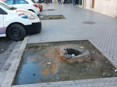 Un coche blanco con la palabra "NISE" en el lado está aparcado a un costado de una calle adoquinada. Hay una fosa en el suelo cerca del coche, con agua y basura alrededor. La imagen sugiere un problema de mantenimiento en la calle o el área circundante.