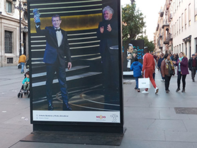 Un hombre en traje azul y sombrero se muestra en un cartel publicitario grande. Detrás de él, una calle con edificios y gente caminando.