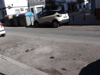 Vista de una calle con dos vehículos estacionados, uno blanco y otro azul. Hay contenedores de basura al lado del camino y una calle con algunas manchas en el asfalto. La imagen muestra un entorno urbano con edificios y una calle vacía.