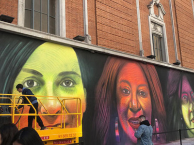 Mural de grandes rostros en un edificio, con personas trabajando en el mural.