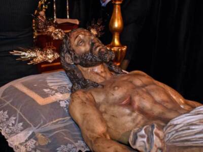 Una estatua de madera tallada detalladamente, con un rostro expresivo y una cabeza con detalles en relieve. La estatua está sobre un tapete de seda con bordados florales, y a su lado hay una figura vestida en negro. La escena parece ser parte de un evento ceremonial o religioso, con una iluminación suave que enfoca en la estatua.