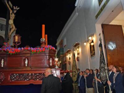En una iglesia, un grupo de personas observa una imagen religiosa. En el centro, hay un altar con flores rojas y un crucifijo en la parte superior. A la derecha, una figura con un manto negro y guantes blancos sostiene un bastón. La pared tiene marcos de fotos y una puerta con un espejo.