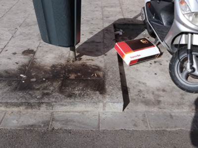 Una caja de cigarrillos descansa sobre el suelo, cerca de una motocicleta aparcada en la acera. La imagen muestra un detalle urbano con elementos cotidianos y una atención a los detalles.