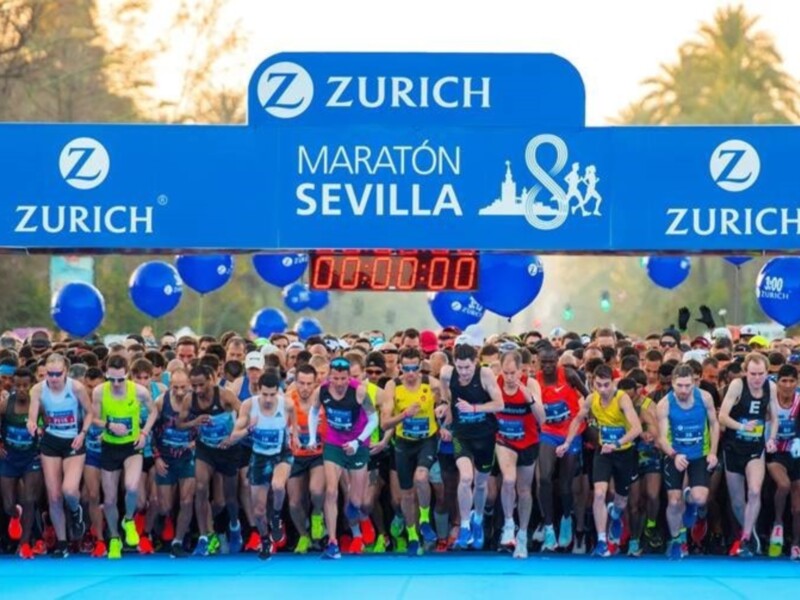 Maratón de Sevilla 2023 con corredores en la línea de salida. Bandera de Zurich y escudo del evento.