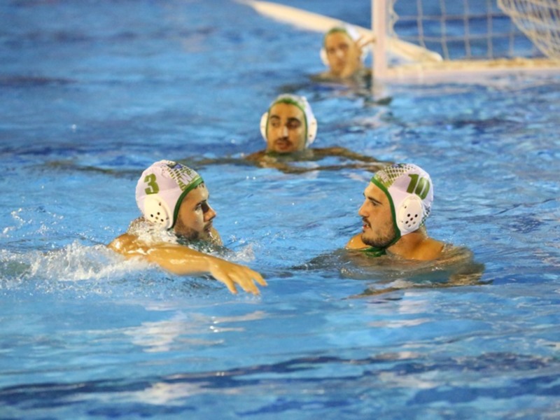 Jugadores de waterpolo en acción, con cascos y ropa deportiva, nadando y jugando en un estadio de agua.