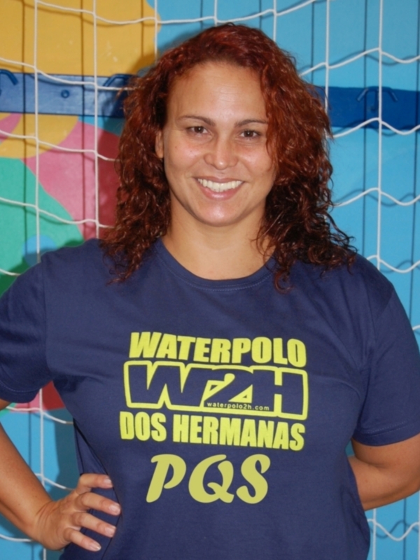Una mujer con cabello rojo largo, sonriendo y posando frente a una valla de portería de fútbol. Lleva una camiseta azul con letras en amarillo y verde que dicen "Waterpolo DOS HERMANAS PQS". El fondo muestra un mural de colores brillantes.