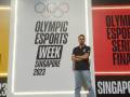 Un hombre con camiseta negra y jeans está posando frente a un panel de anuncios que dice "Olympic Esports Week Series Finals 2023".