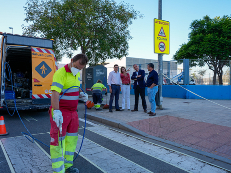 Trabajador de limpieza en uniforme amarillo y rojo, con una máscara facial blanca, se prepara para limpiar el pavimento. En la imagen, hay un poste de señalización amarillo con una advertencia sobre velocidad y un coche de limpieza en el fondo. La escena se desarrolla en una calle con árboles y un poste de luz azul.