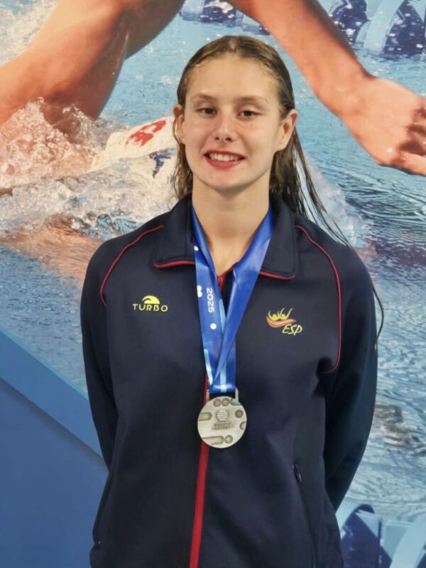 Una nadadora posa con una medalla de plata en un evento deportivo, con otra nadadora en el fondo.