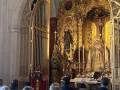 Interior de una iglesia con arcos dorados y estatuas religiosas. Personas en oración frente a un altar ricamente decorado con flores y velas.