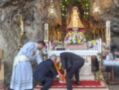 En una cova rocosa, tres personas rezan ante un altar adornado con flores y una imagen religiosa. La escena transmite seriedad y devoción en un ambiente de piedra natural.