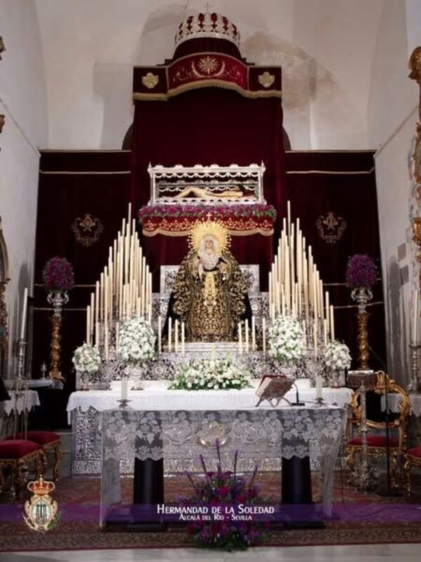 Imagen de una iglesia con un altar central adornado con flores y velas. En el fondo, se puede ver una imagen religiosa en un trono. La imagen está firmada por "Hermanadad de la Soledad" y se encuentra en Alcala de los Gazules, España.