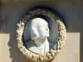 Escultura de mármol en relieve que representa un rostro humano con una corona de laurel, situada en la parte superior derecha de un pedestal de piedra. La imagen muestra una escena histórica o cultural, posiblemente de un monumento o sepulcro.