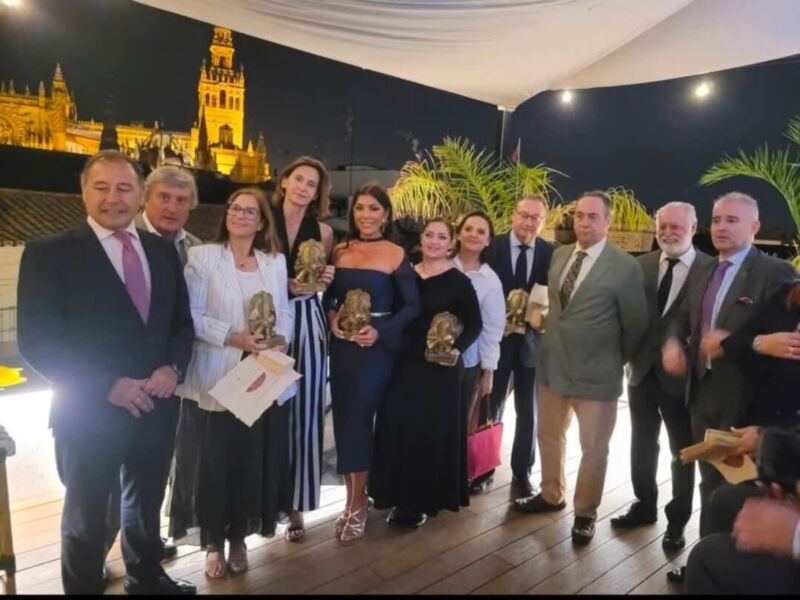 A una grupo de personas posando para una foto en un evento formal. Al fondo se puede ver la famosa Giralda de Sevilla, España. Las personas están vestidas formalmente y algunos portan reconocimientos o premios dorados. La escena parece ser en un ambiente de celebración o gala.