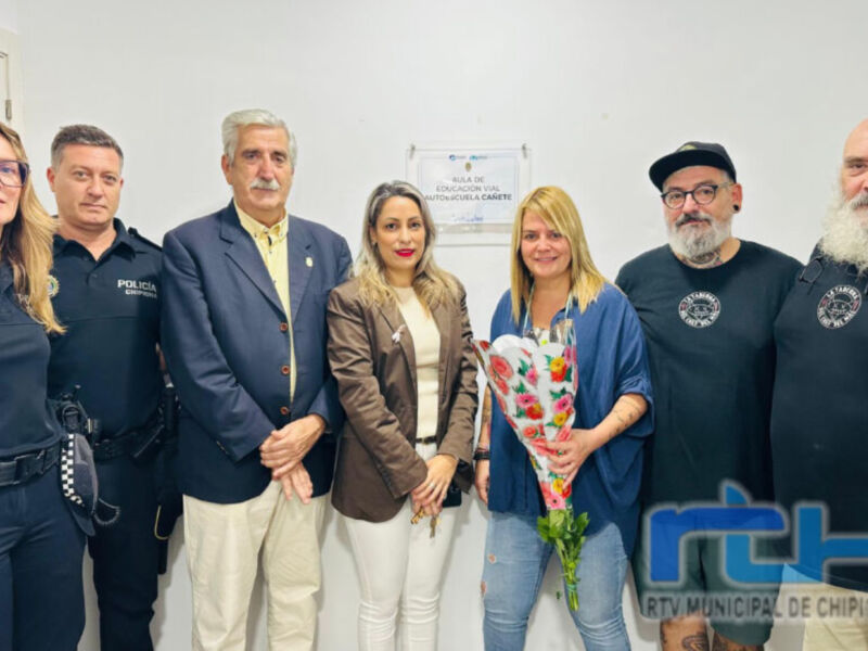 Una imagen de una reunión formal con varias personas en uniforme y civiles. En el centro, una mujer sostiene un ramo de flores, mientras otros individuos observan la reunión. En el fondo, se puede ver un cartel con información sobre una conferencia o evento. La imagen está firmada por "RTV Municipal de Chipiona".