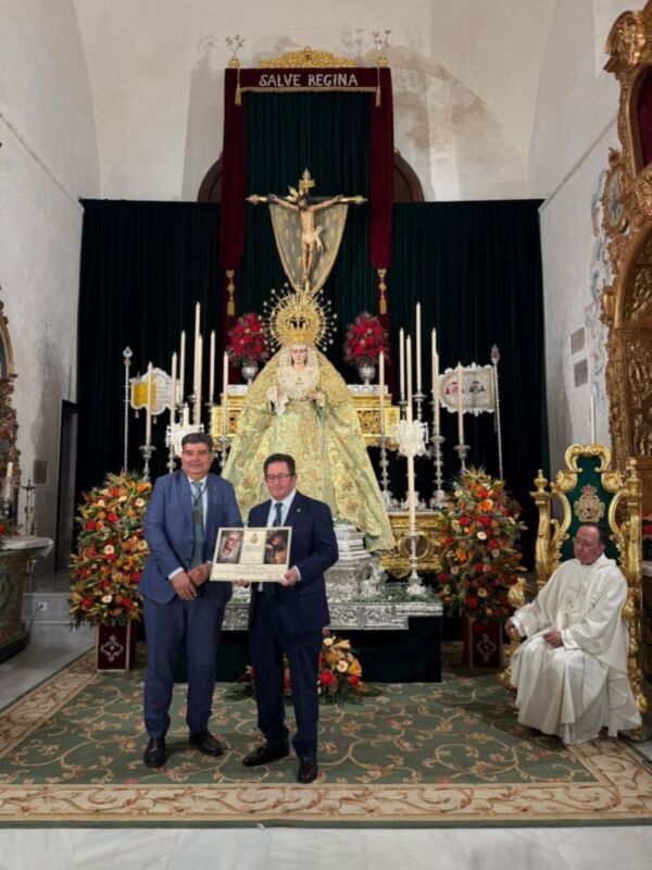 Dos hombres en trajes de negocios sostienen un libro mientras una persona vestida con un hábito religioso se sienta en la derecha. La imagen muestra una escena formal con elementos religiosos y decorativos, incluyendo un altar con una imagen de la Virgen María y el Santo Cristo. La decoración incluye flores y una alfombra con patrones geométricos en el suelo.