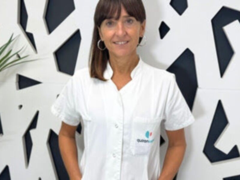 Una mujer con cabello largo y recto, vistiendo una camisa blanca con un logotipo en la manga izquierda. Detrás de ella, una pared decorada con formas geométricas negras y blancas.