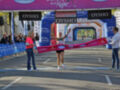 Una corredora femenina celebra su victoria en una carrera de maratón, cruzando la línea de meta con un atleta femenino ayudándola a levantar el banner de victoria.