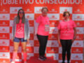 Tres personas posan en un podio rojo con el logo de Santander, celebrando su logro. La persona en el primer lugar sostiene un trofeo y está vestida con una camiseta rosa. La segunda persona, en el segundo lugar, también lleva una camiseta rosa y pantalones negros. La tercera persona, en el tercer lugar, lleva una camiseta rosa y pantalones negros. El fondo es un muro de anuncios rojos con el texto "¡OBJETIVO CONSEGUIDO!".