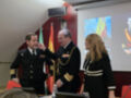 Tres personas en uniforme militar, dos hombres y una mujer, se encuentran en un evento formal. La imagen muestra a los individuos hablando y gesturando, con una pantalla proyectada en el fondo. La escena se desarrolla en un entorno interior, posiblemente una sala de conferencias o auditorio.