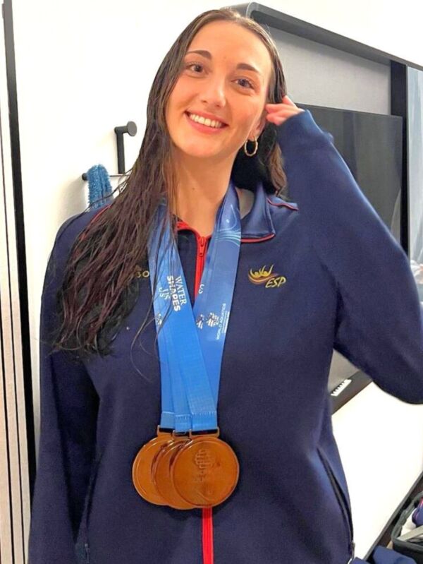 Una mujer sonríe mientras sostiene una medalla de oro en la cintura, vestida con un uniforme azul marino que lleva el logo de España.